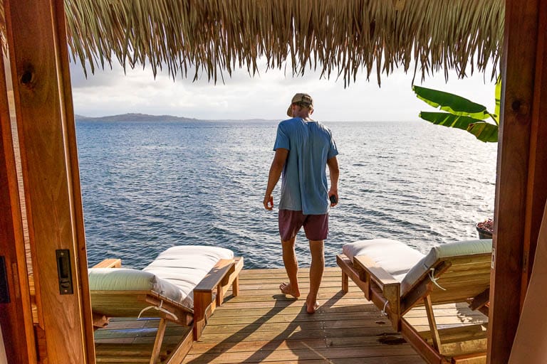 A man walks in to see the water of an overwater bungalow during sunset.