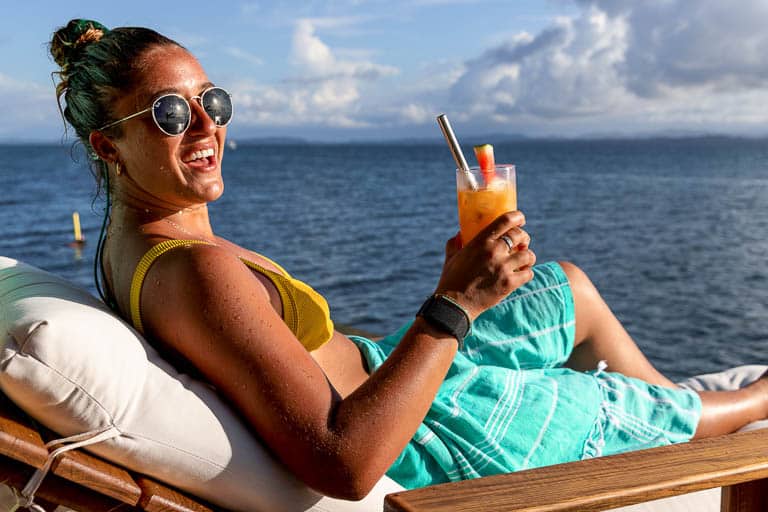 A guest of Sol Bungalows in a yellow bikini smiles while drinking an orange and yellow tropical drink.