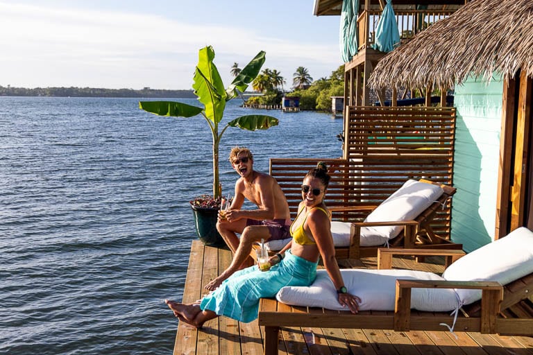 A couple smile at the camera while relaxing on the chaise lounge chairs on the deck of an overwater bungalow in panama.