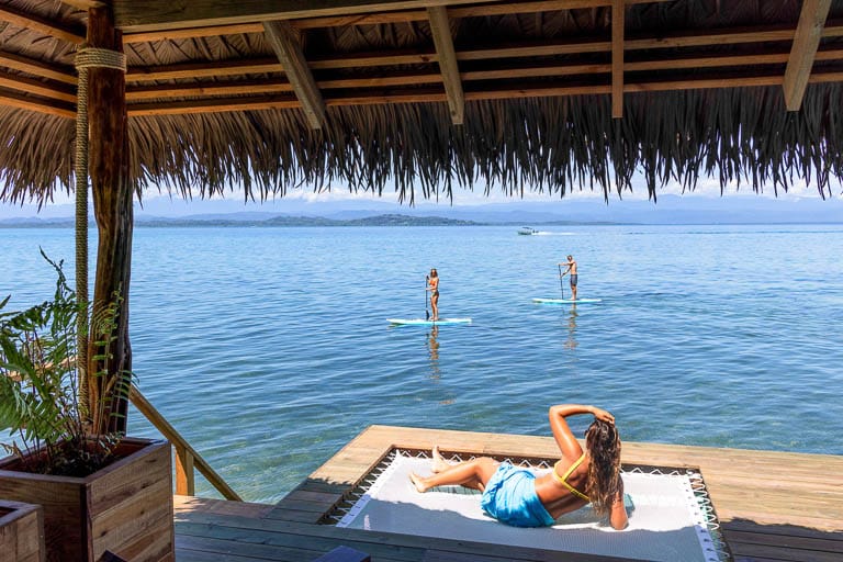 A woman lays in the hammock of an overwater bungalow while two paddle boarders cruise by in the distance.