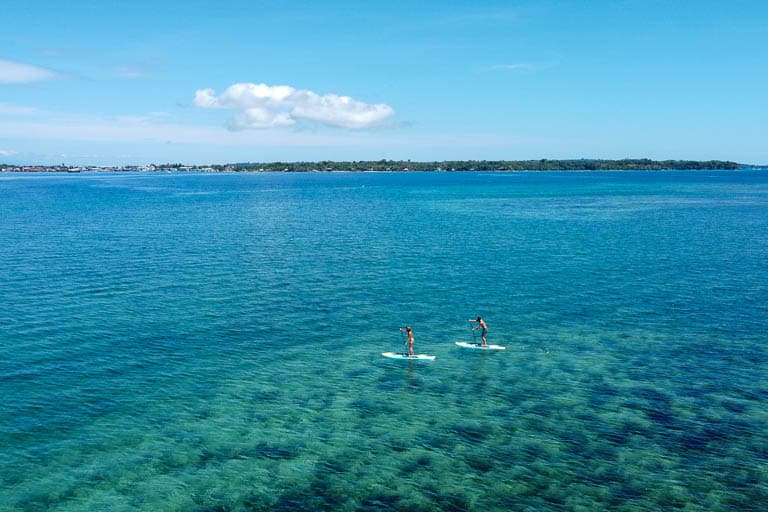 Clear caribbean water dominates the scene where two paddle boarders cruise over a shallow reef.