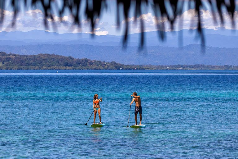 A beautiful blue caribbean sea is the backdrop to two paddle boarders.