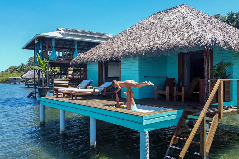 Two women in bikinis practice acro yoga on a catamaran net in front of the Sol Bungalows overwater bungalows.