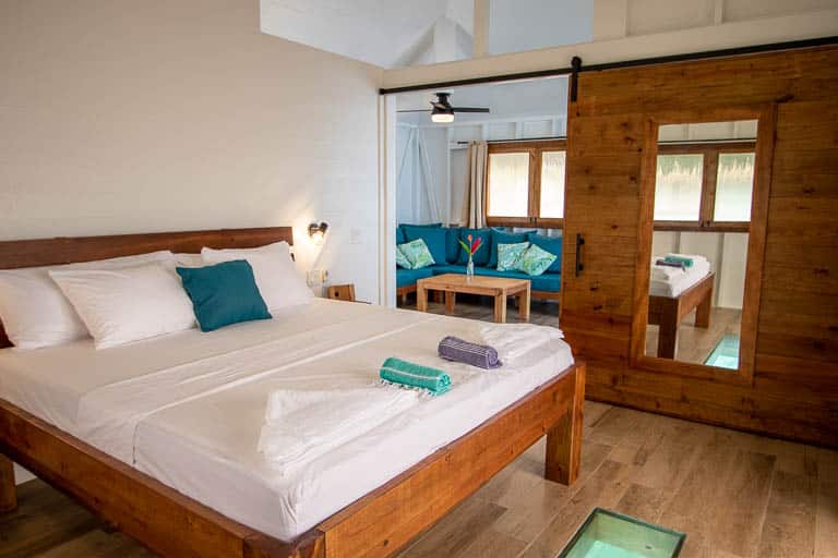 The interior of the overwater bungalows at Sol Bungalows which have king size beds, glass floors, sliding walls with a mirror, ceiling fans, and white walls with high ceilings.