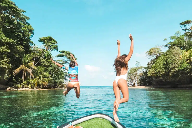 Two women in bikinis jump off the front of a panga boat into the blue water of Bocas del Toro at Bird Island