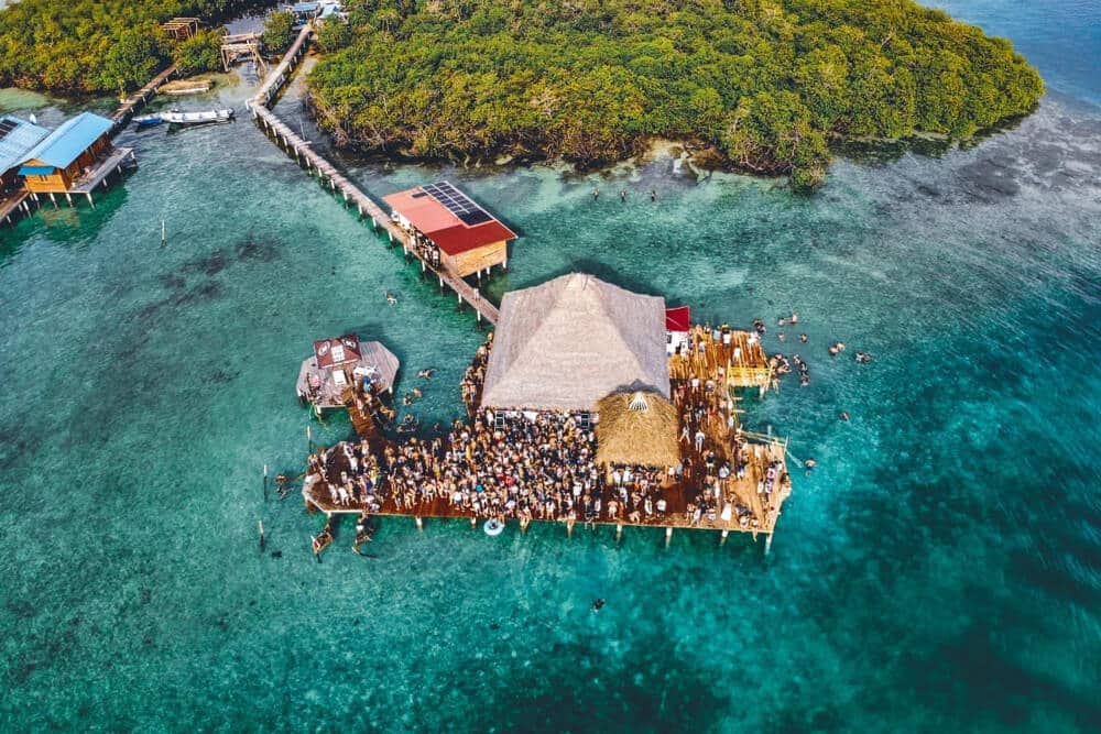 Filthy Friday party at the overwater restaurant called the blue coconut on isla solarte in bocas del toro panama. The view is from a drone, showing clear blue water, a thatch roofed restaurant, and the outside deck full of party goers.